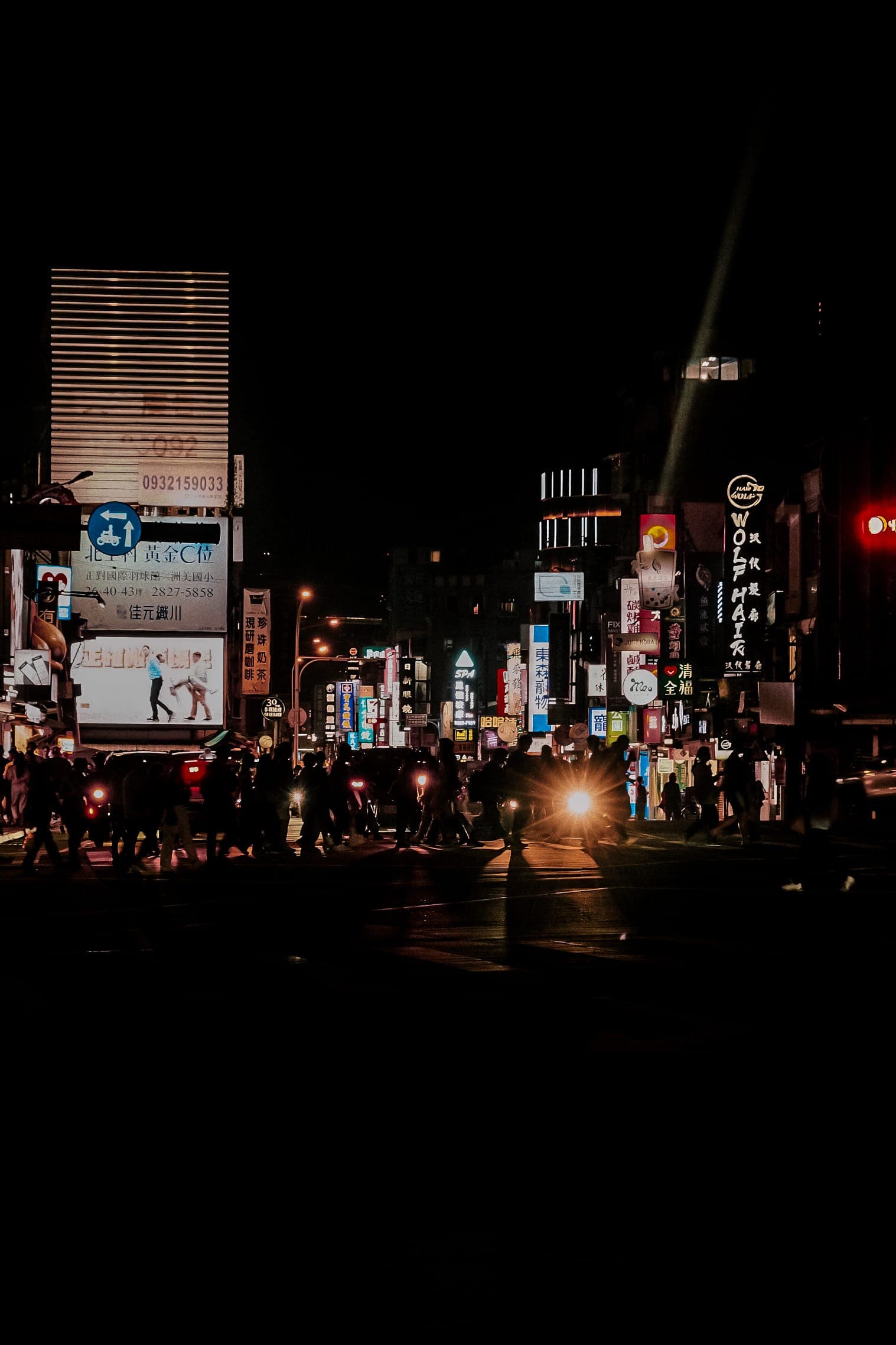 Pedestrians crossing a Taipei intersection late at night, headlights flaring.