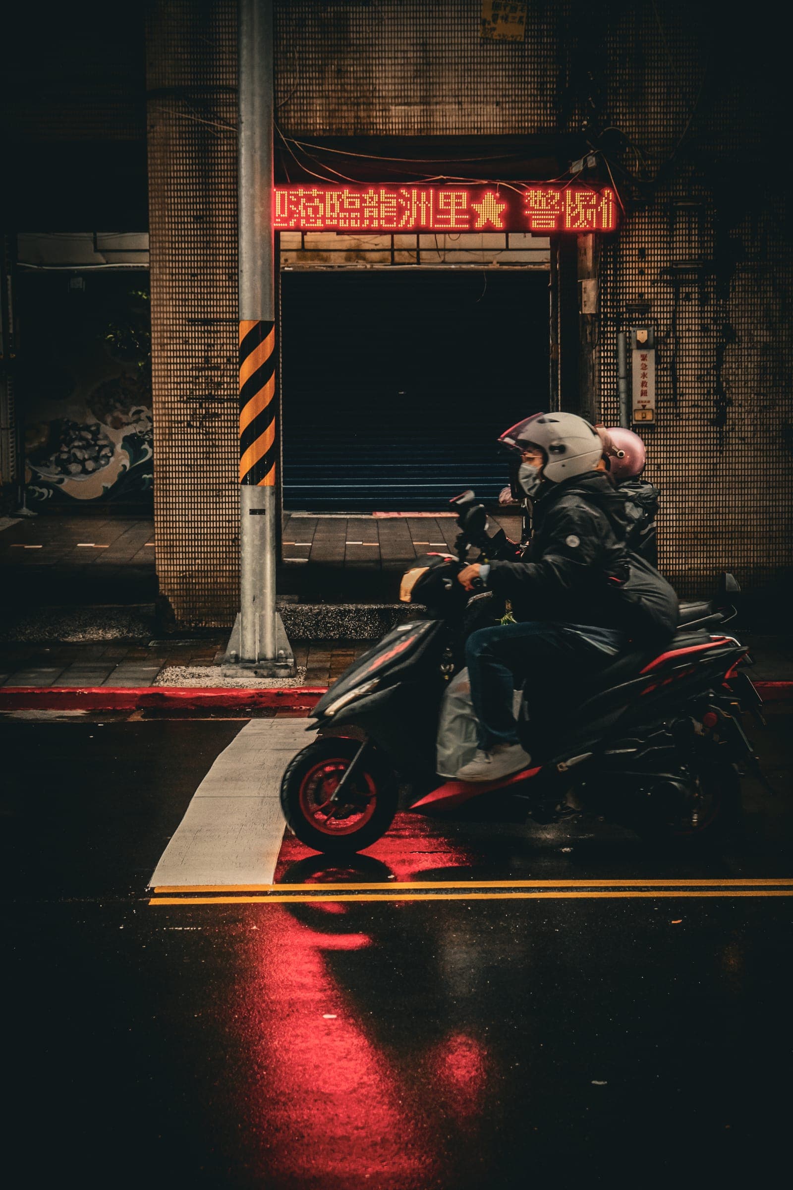 A scooter rider waiting under a red neon sign on wet Taipei pavement.