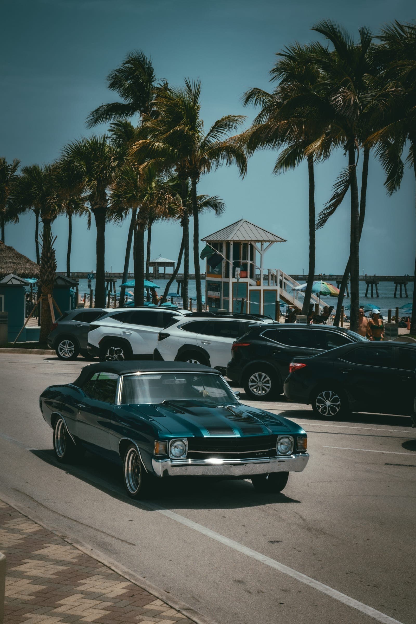 A teal 1972 Chevrolet Chevelle convertible with black stripes rolling past beachfront palms, a lifeguard tower and pier behind.