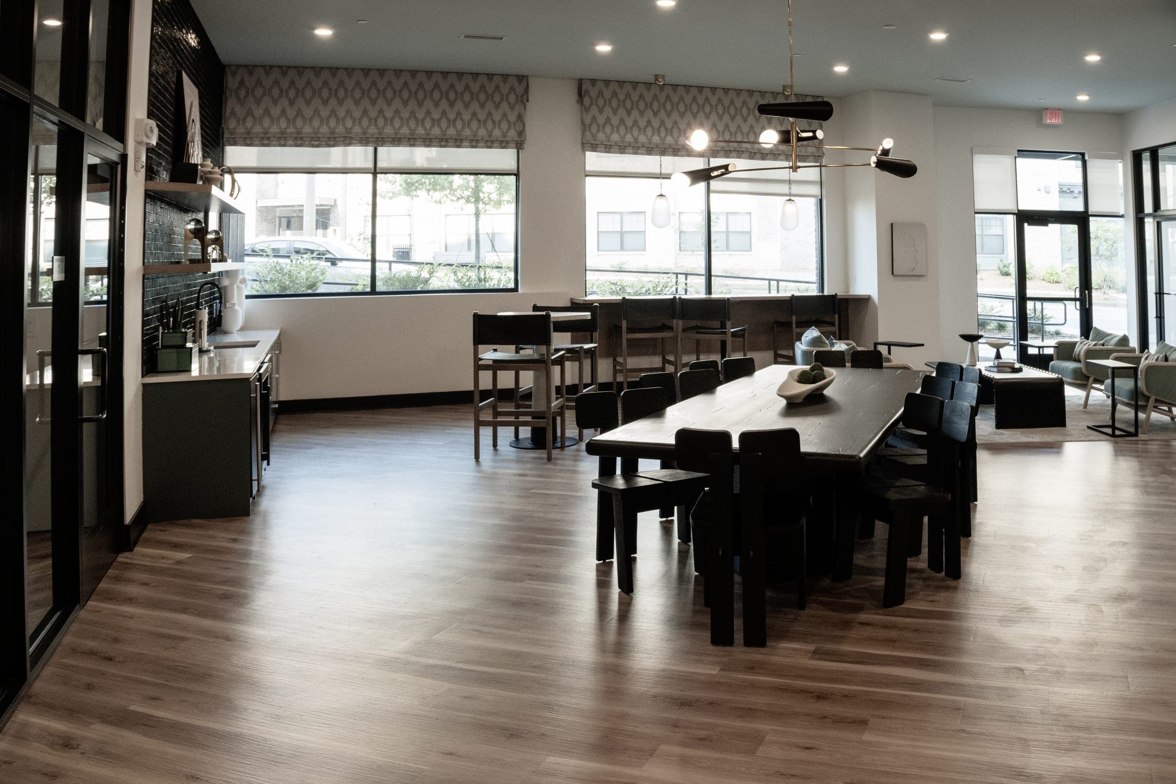 Community clubhouse dining area with a long black table, patterned roman shades, and a brass mobile chandelier.