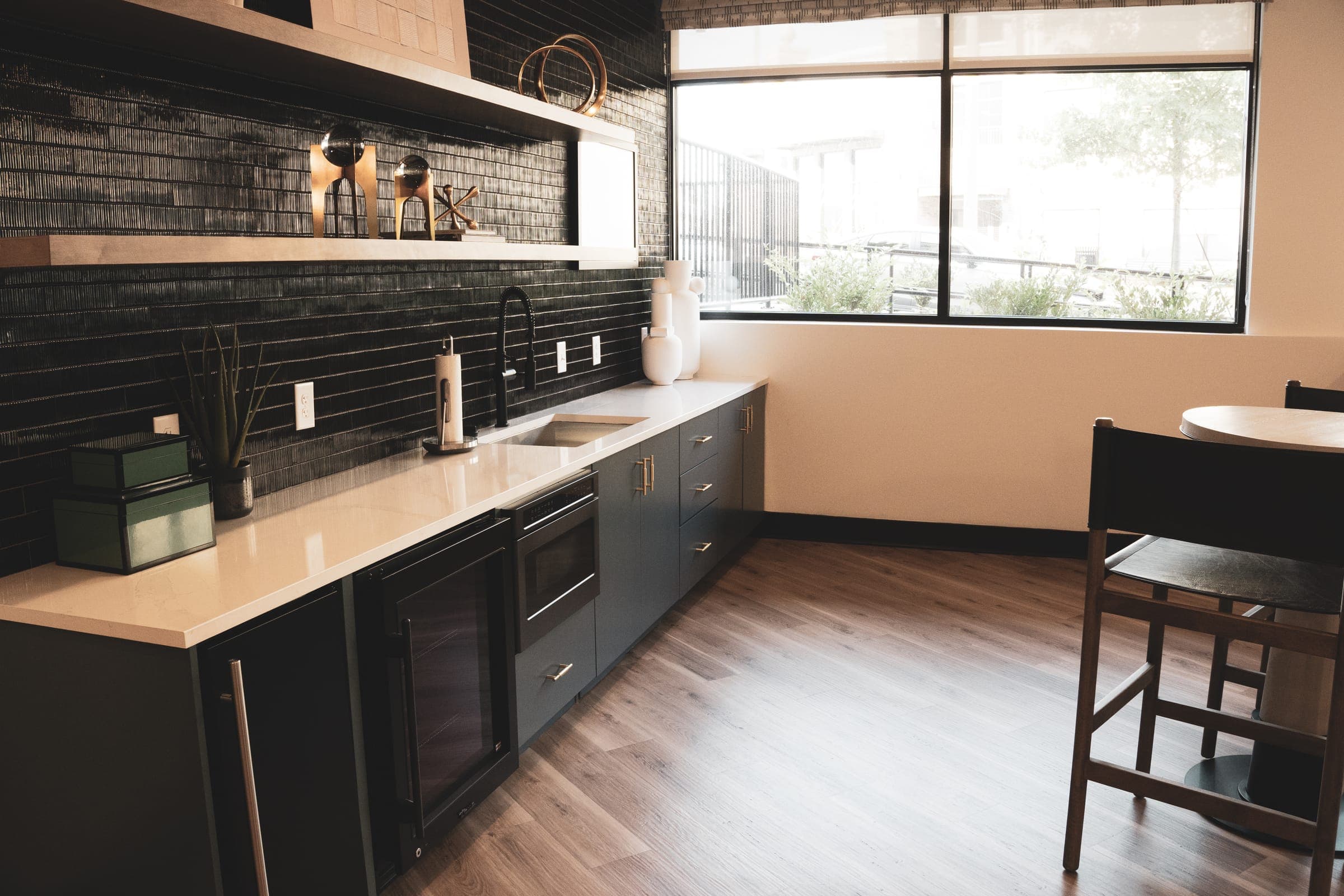 Amenity lounge kitchenette with glossy black tile backsplash, white quartz counters, and a wine fridge under warm window light.